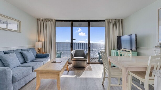 A modern living room with a sofa, a coffee table, a TV, and a balcony with a view of the sea. - 0609 Oceanfront Escape by Atlantic Towers (Carolina Beach)