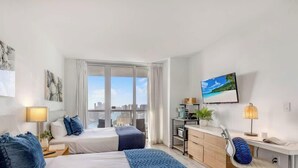 A hotel room with a bed, bedside table, lamp, and a view of the city through large windows.