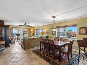 A spacious living area with a dining table, chairs, and a sofa. There is a balcony with a view, and the room has a ceiling fan. - Regency Towers #408 East | Gulf Views, Pool! (Pensacola Beach)