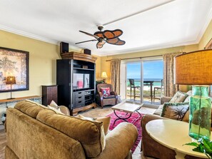 A living room with a sofa, a television, a ceiling fan, and a view of the ocean through a sliding glass door.