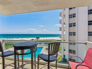 A balcony with a view of a beach and ocean, a pool, and outdoor furniture.