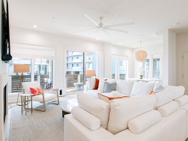 A modern living room with a white sofa, a coffee table, and a floor lamp. There is a balcony with a view of buildings.