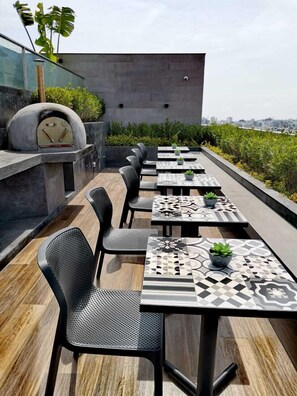 Una terraza en la azotea con mobiliario exterior, un horno de pizza y vistas de la ciudad.