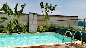 A rooftop swimming pool with a wooden deck, surrounded by large tropical plants and a modern building in the background.