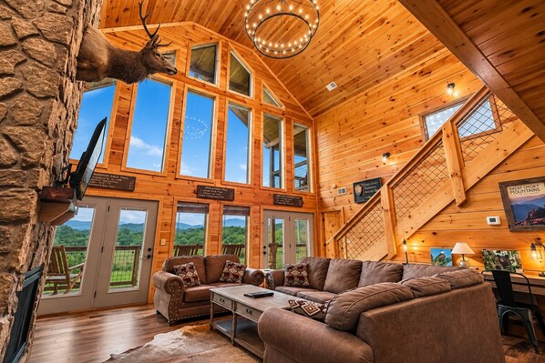 A log cabin interior with a large wooden ceiling, multiple windows, a fireplace, and a stone wall.
