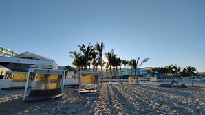A beach with sun loungers and umbrellas, palm trees, and a building in the background.