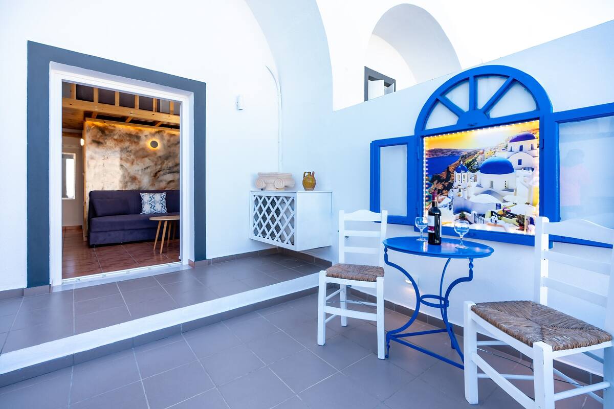 Un intérieur moderne avec un coin salon comprenant des chaises blanches et une table bleue, un mur décoratif orné d’un tableau, et une porte-fenêtre donnant sur une autre pièce.