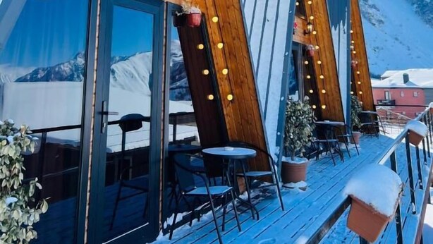 A modern, triangular-roofed building with a wooden deck and outdoor seating area, set against a snowy mountain backdrop. - PYRAMID KAZBEGI (Kazbegi)