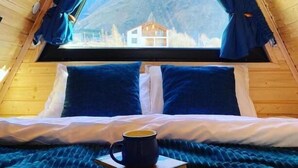 A cozy, triangular wooden room with a bed, a cup of coffee, and a view of a mountain and a house. - PYRAMID KAZBEGI (Kazbegi)