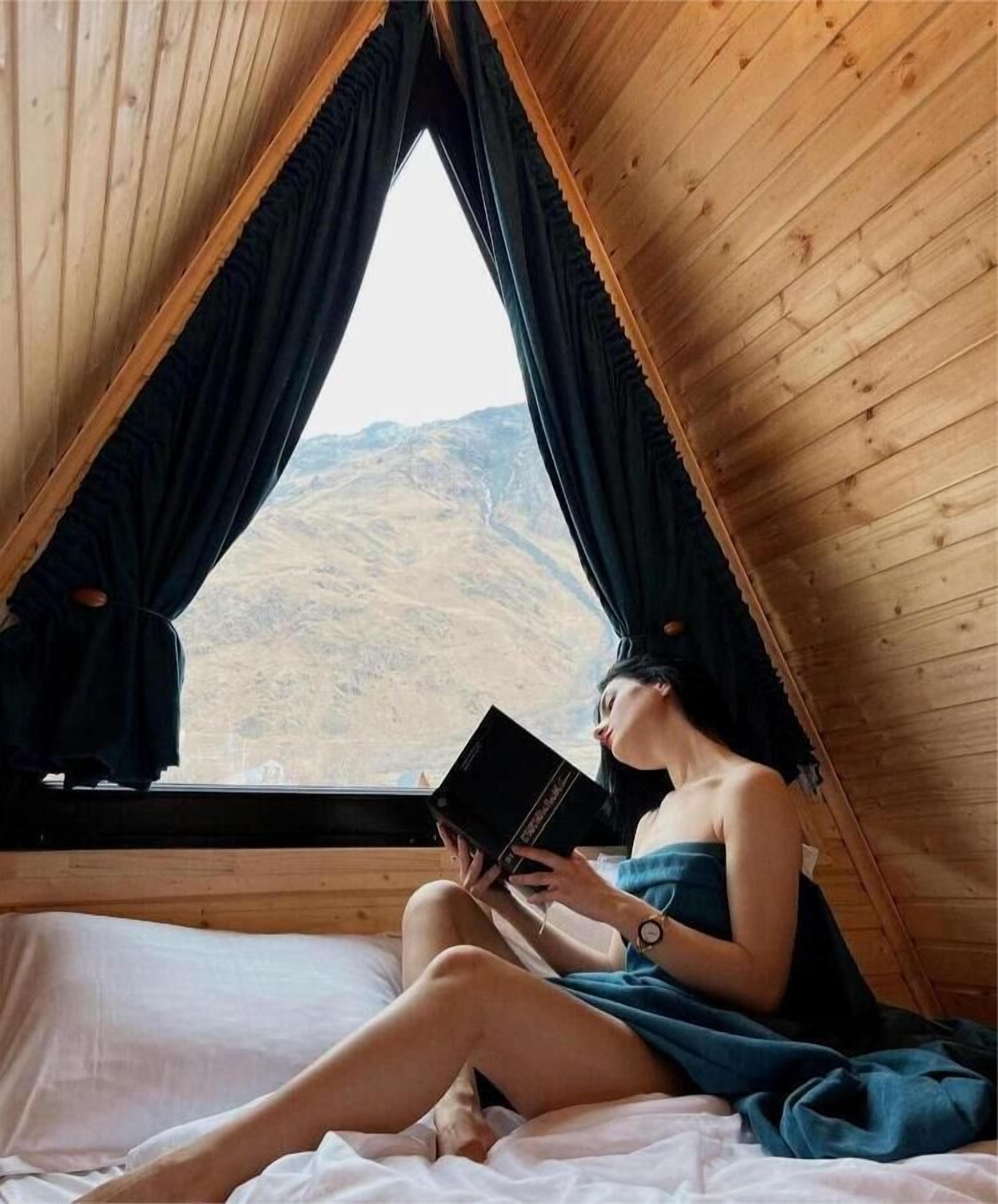 Uma pessoa sentada em uma cama em um quarto de madeira, com vista para uma paisagem montanhosa pela janela.