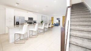 Eine moderne Küche mit weißen Schränken, einer Granit Arbeitsplatte und Edelstahlgeräten. Im Essbereich befinden sich weiße Barhocker und ein blaues Sofa.