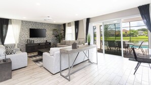 Ein modernes Wohnzimmer mit einem Flachbildfernseher, grauen Sofas, einem Couchtisch und Blick auf einen Swimmingpool durch große Glasüren.