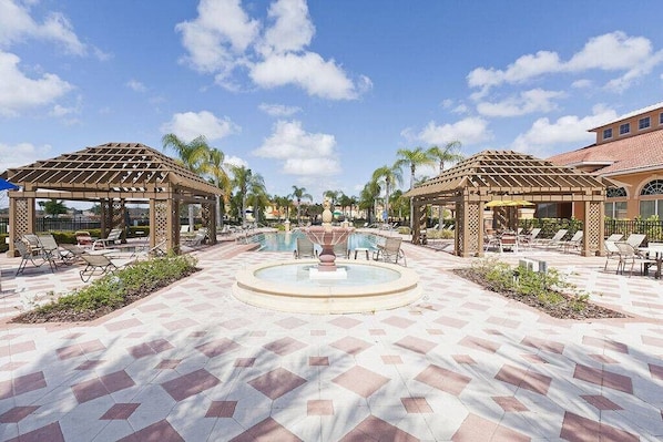 A resort pool area with two wooden gazebos, lounge chairs, and a central fountain.