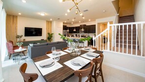 A modern dining area with a wooden table set for four, a kitchen in the background, and a staircase leading to an upper floor.