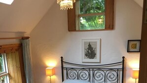 Une chambre avec un plafond en pente, un lit avec un cadre en métal, deux lampes de chevet, un lustre, une fenêtre donnant sur des arbres et un tableau encadré au mur.