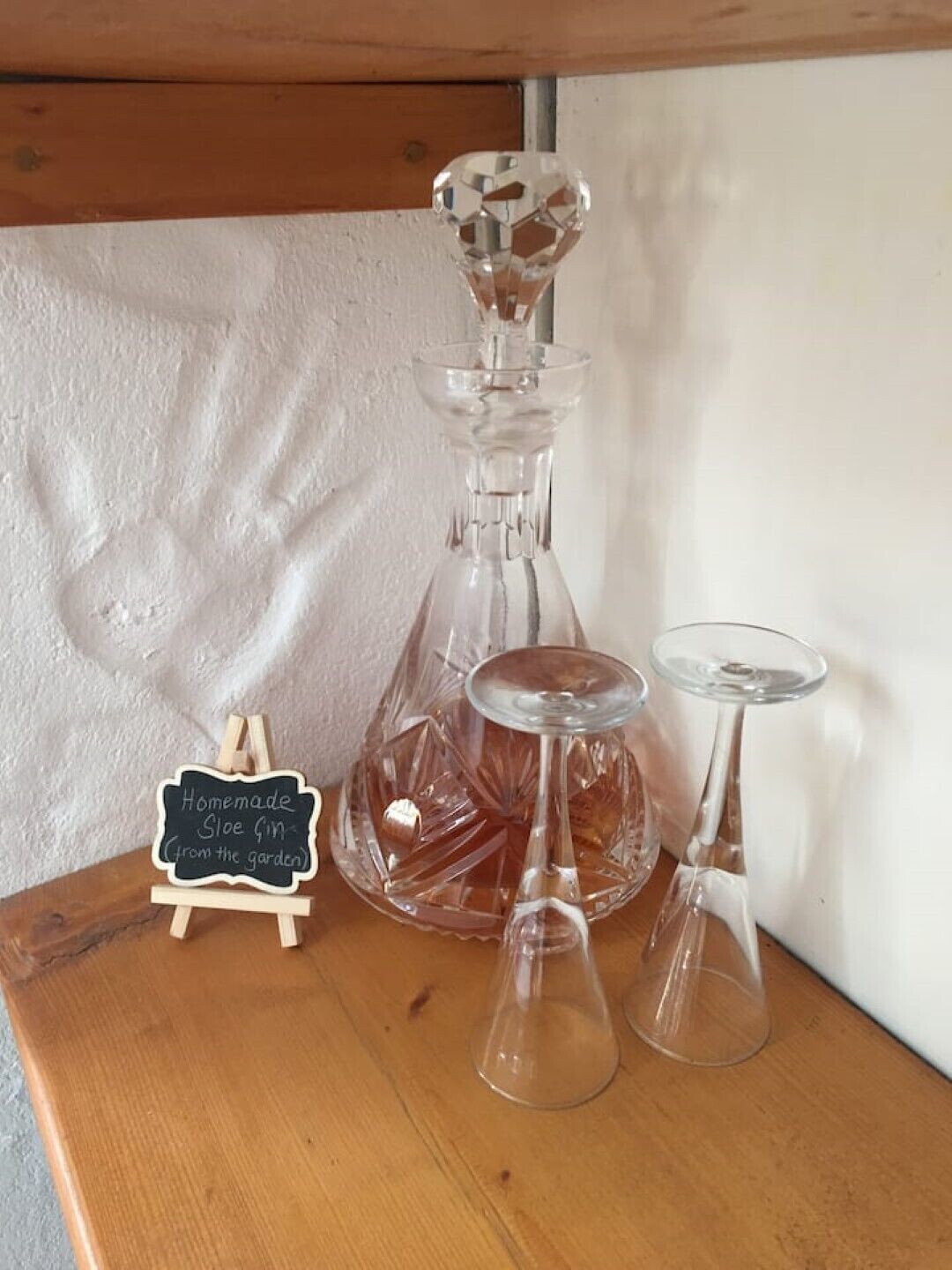 Une table en bois, sur laquelle se trouvent une carafe en verre, un petit tableau noir et deux verres à vin.