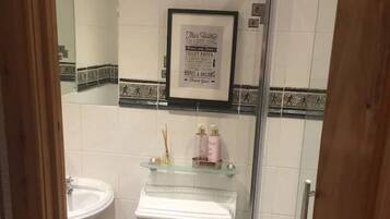 Une petite salle de bain comprenant des toilettes, un lavabo et une douche. Un panneau encadré est accroché au mur, et des articles de toilette sont posés sur le lavabo.