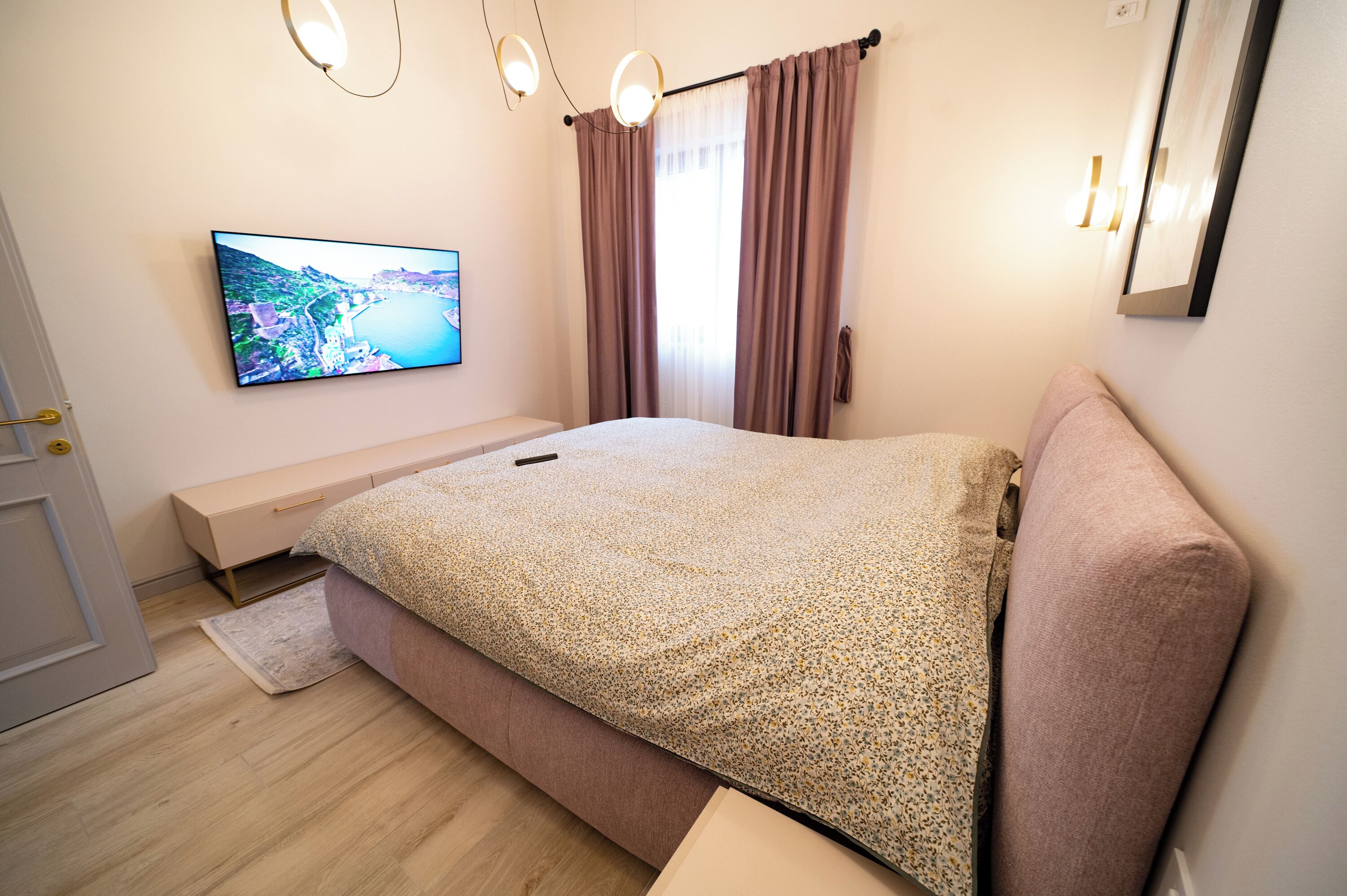 Une chambre à coucher avec un lit, un téléviseur fixé au mur, un banc et une fenêtre avec des rideaux.