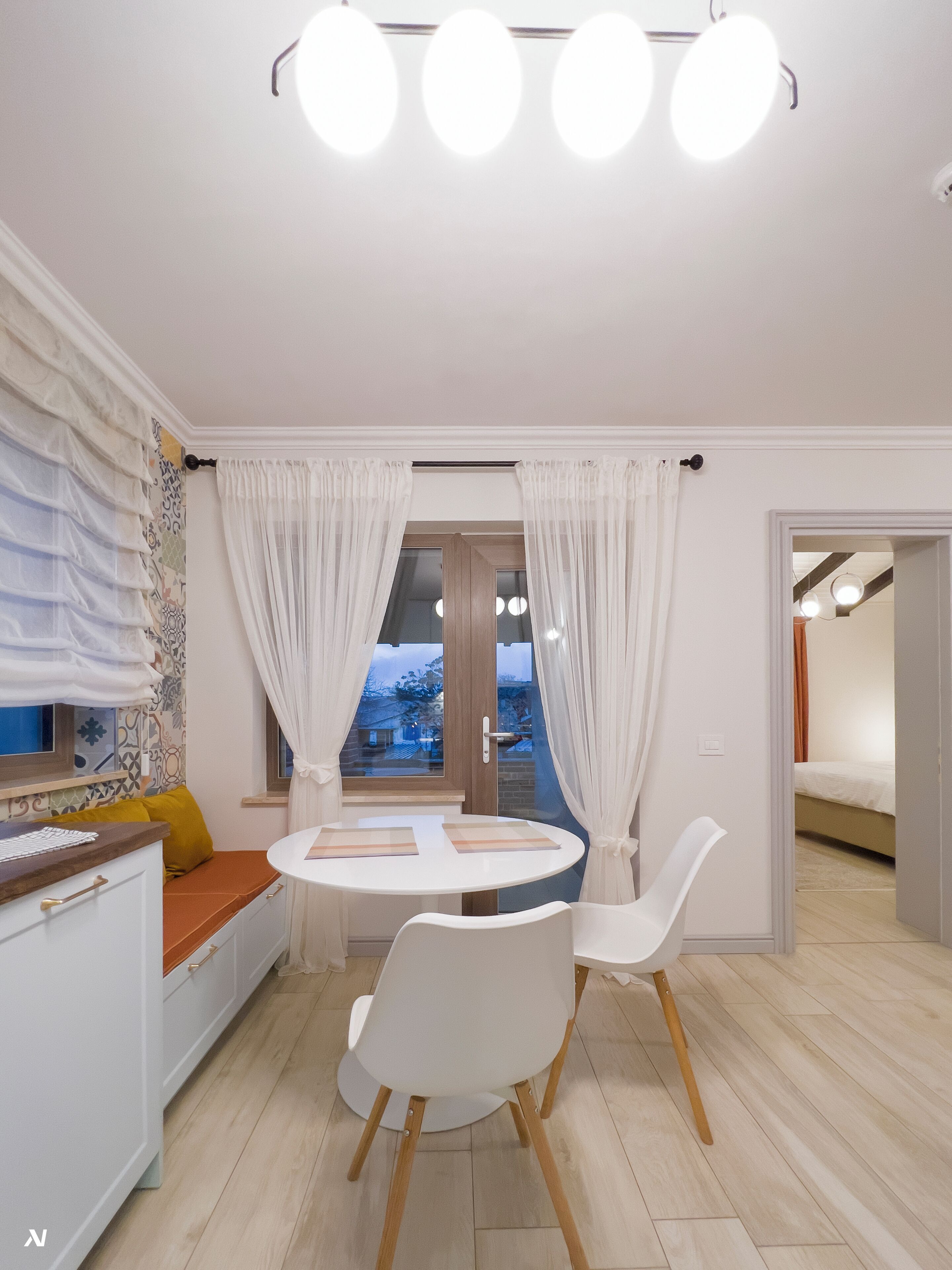 Eine moderne Küche mit weißen Schränken, einem runden Esstisch und zwei weißen Stühlen. Ein Fenster mit Vorhängen bietet Blick ins Freie, und eine Tür führt in einen weiteren Raum.