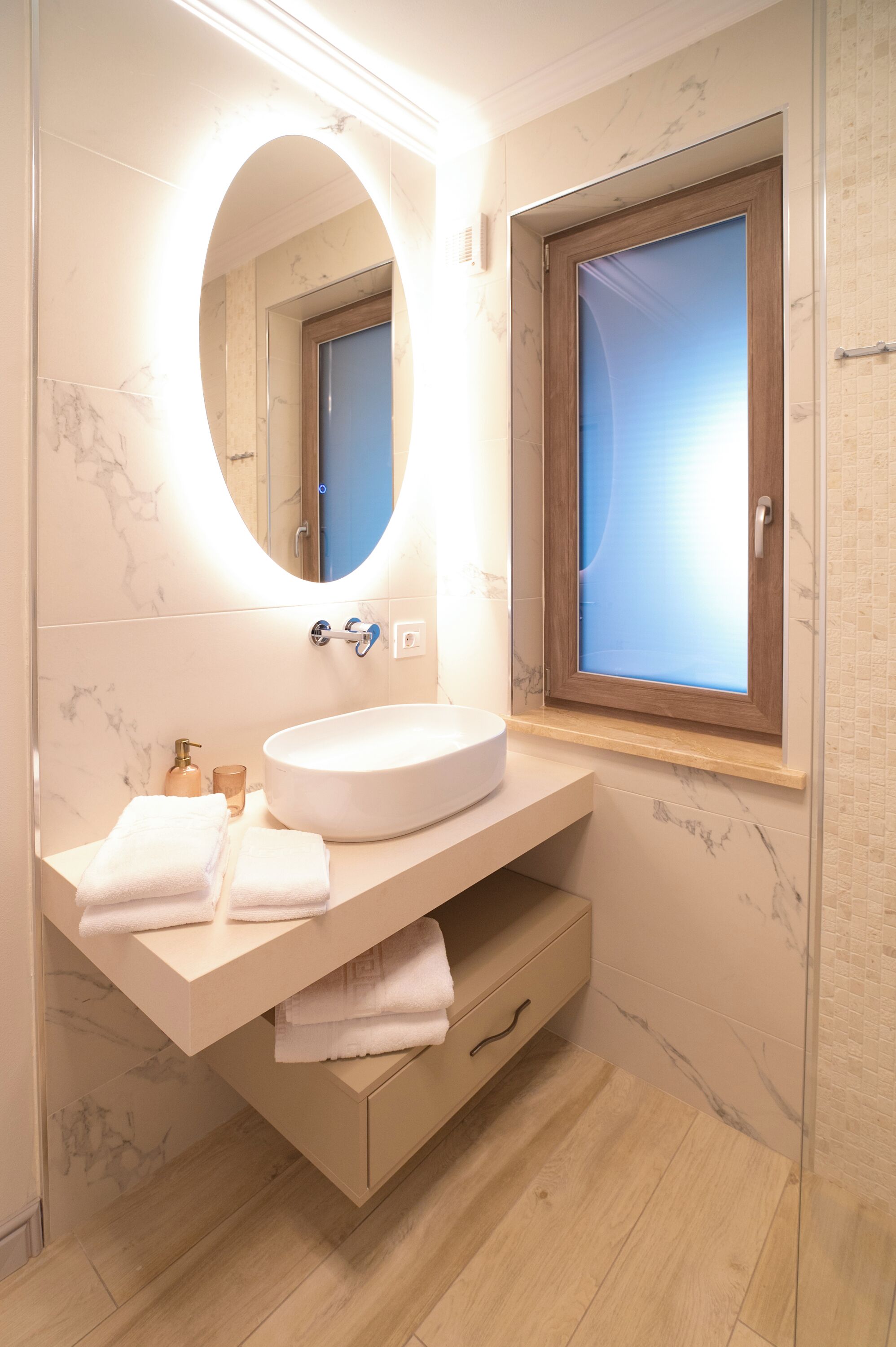 A bathroom with a marble-tiled wall, a white sink, a mirror, and a window with a wooden frame.