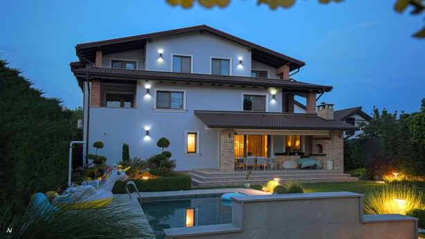 A two-story house with a swimming pool, outdoor seating, and well-lit windows.