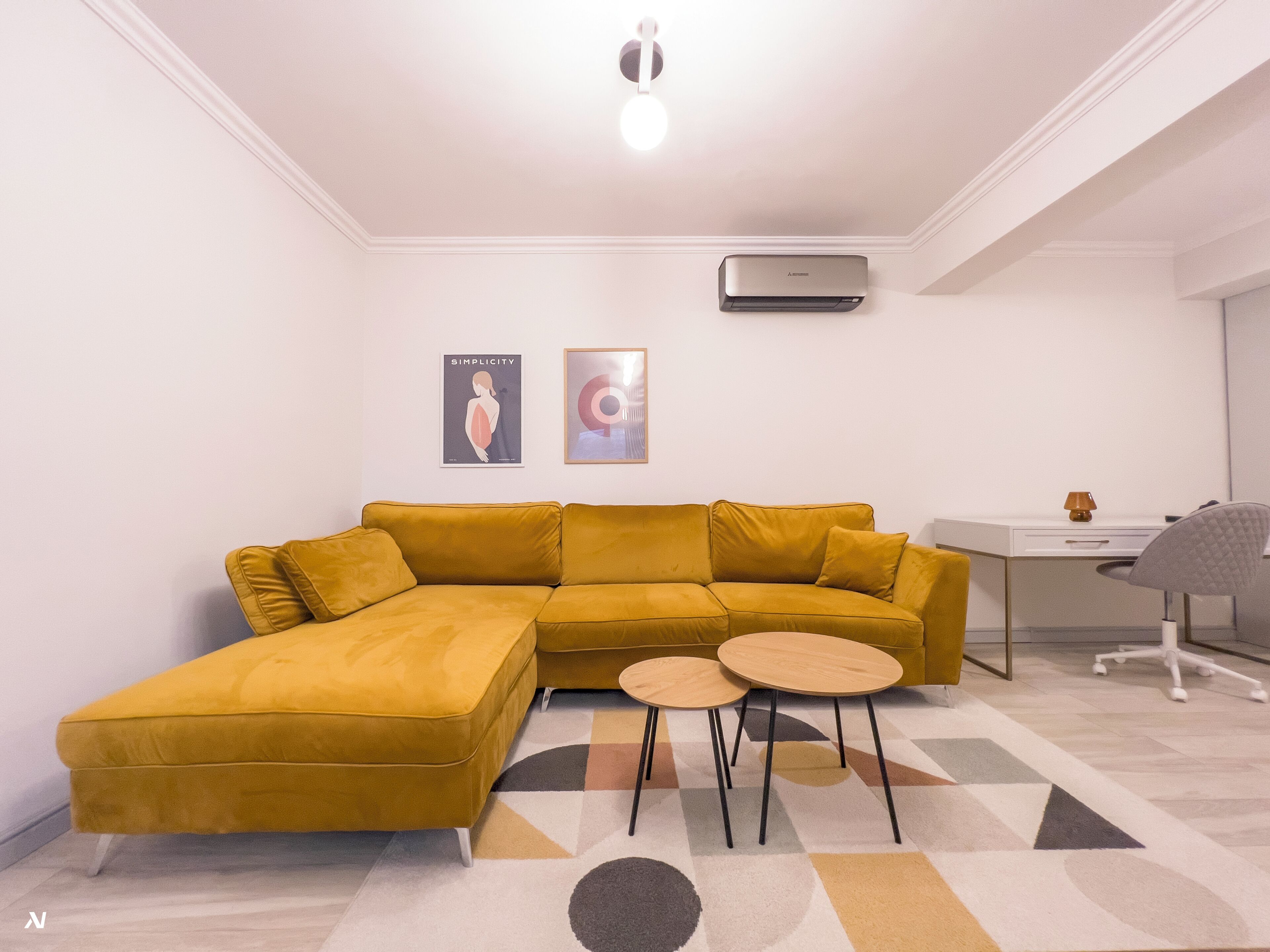 A modern living room with a mustard-colored sectional sofa, a small round table, and a desk with a chair. There are two framed artworks on the wall and an air conditioning unit above the sofa.