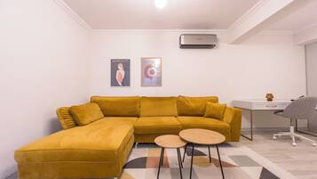 A modern living room with a mustard-colored sectional sofa, a small round table, and a desk with a chair. There are two framed artworks on the wall and an air conditioning unit above the sofa.