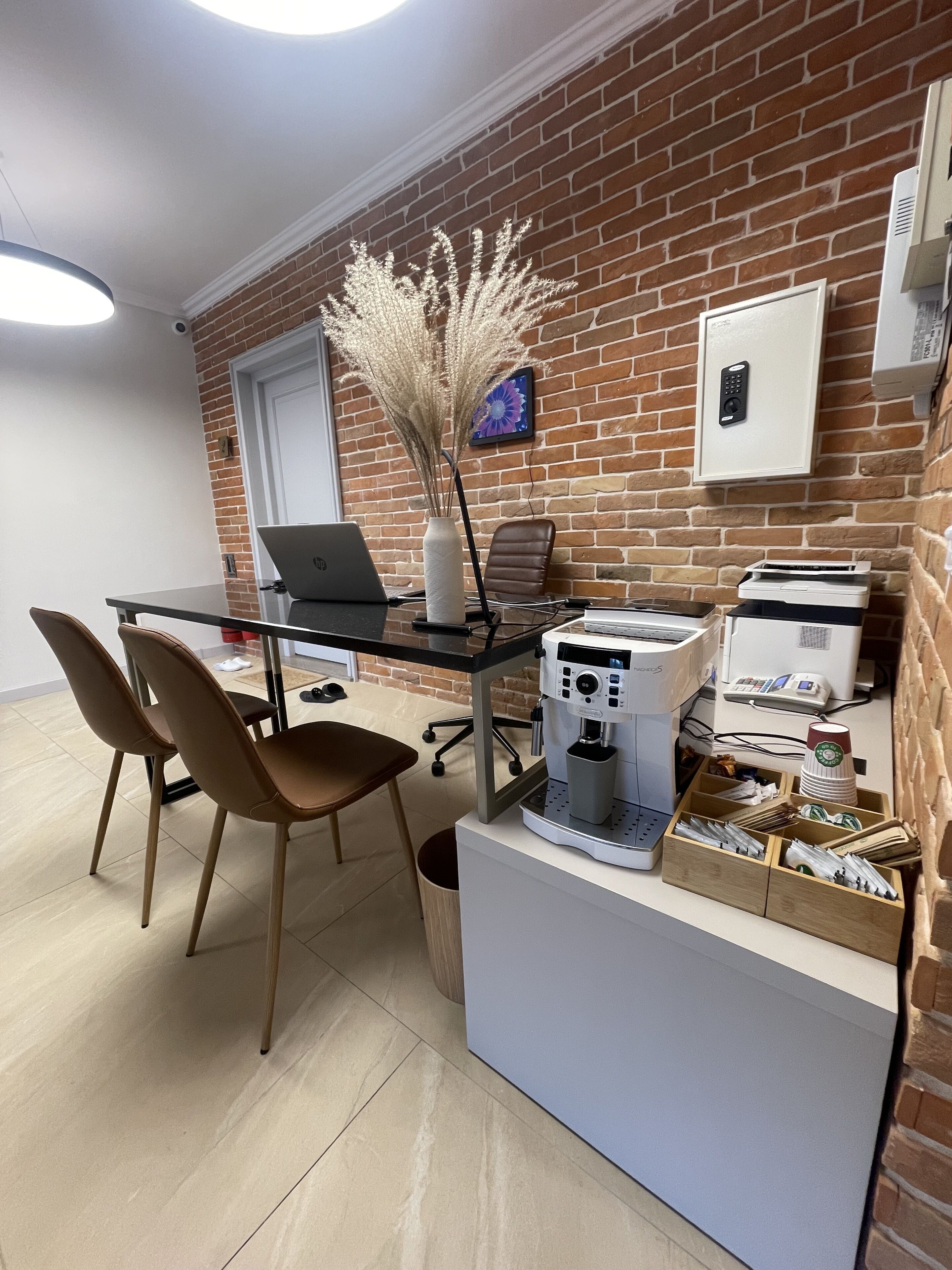 A modern office space with a brick wall, a glass desk, two brown chairs, a laptop, a coffee machine, and a printer.