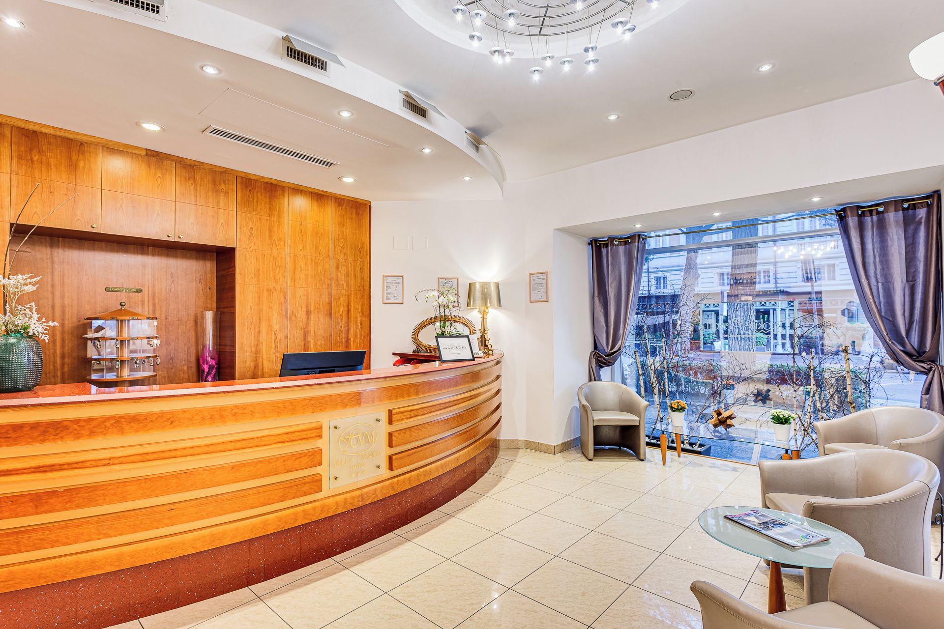 A hotel reception area with a wooden reception desk, a computer, and a seating area with two chairs and a small table.