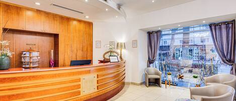 A hotel reception area with a wooden reception desk, a computer, and a seating area with two chairs and a small table.
