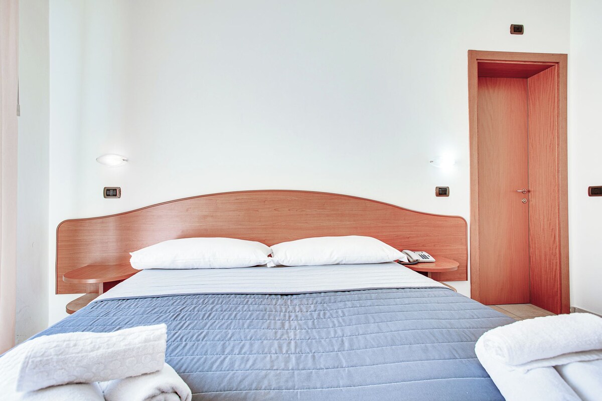 Un lit bien fait, avec une tête de lit en bois, des oreillers blancs et une couverture bleue. On aperçoit une table de chevet en bois sur laquelle se trouvent un téléphone et une petite lampe. Une porte en bois est visible sur la droite.
