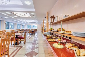 A spacious dining area with multiple tables and chairs, a buffet counter with various food items, and large windows allowing natural light. - Villa Maria (Cesenatico)