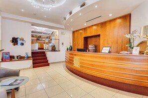 A hotel reception area with a curved wooden reception desk, a staircase leading to another floor, and a glass coffee table with magazines. - Villa Maria (Cesenatico)