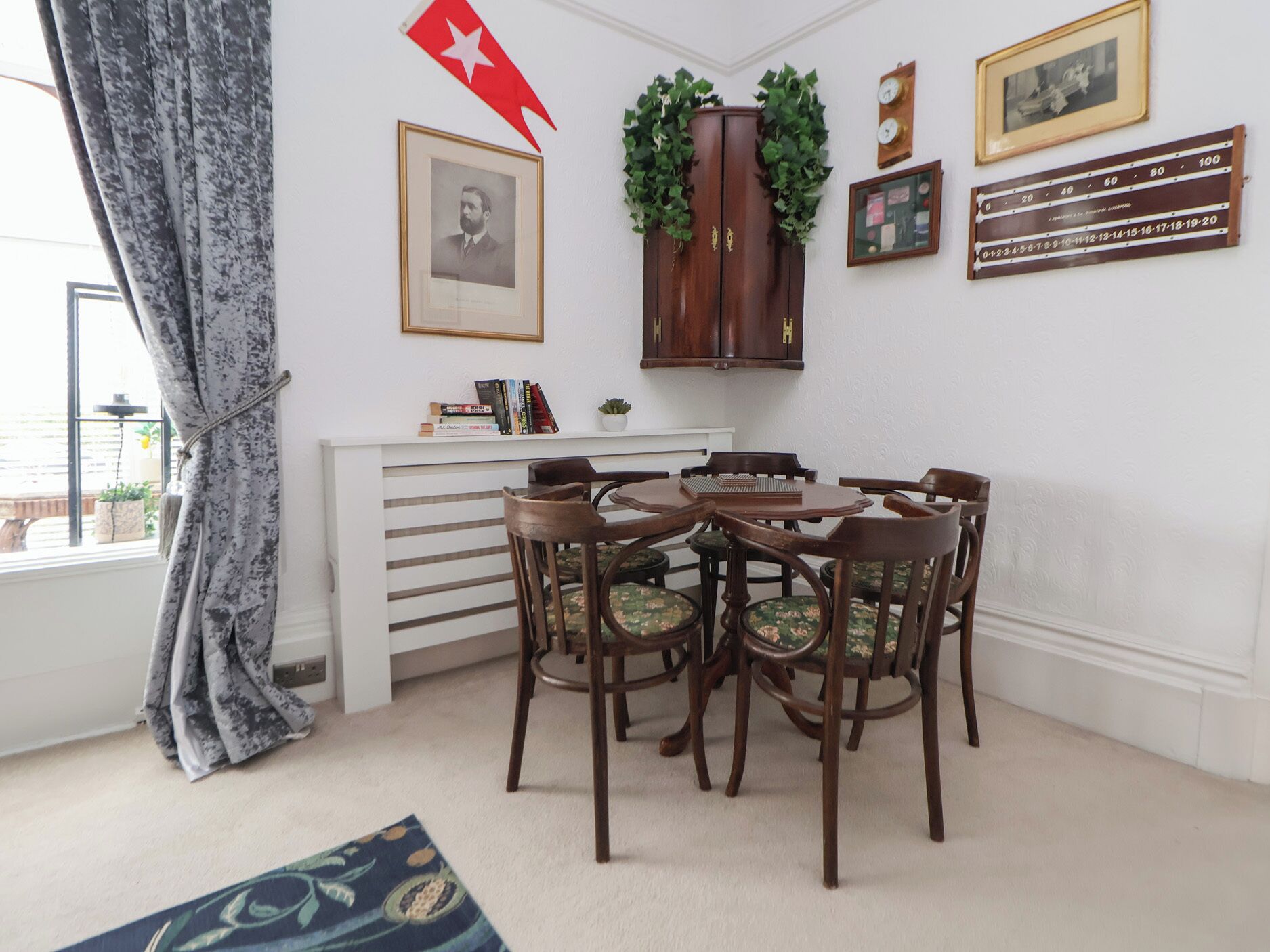 A dining area with a round table and six chairs, a bookshelf with books, a framed portrait, a clock, and a red flag.
