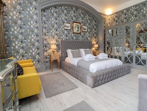 A bedroom with a large bed, a nightstand, a lamp, a mirror, and a yellow chair. - Ismay Billiard Room Apartment - Titanic Interest Beach View (Liverpool)