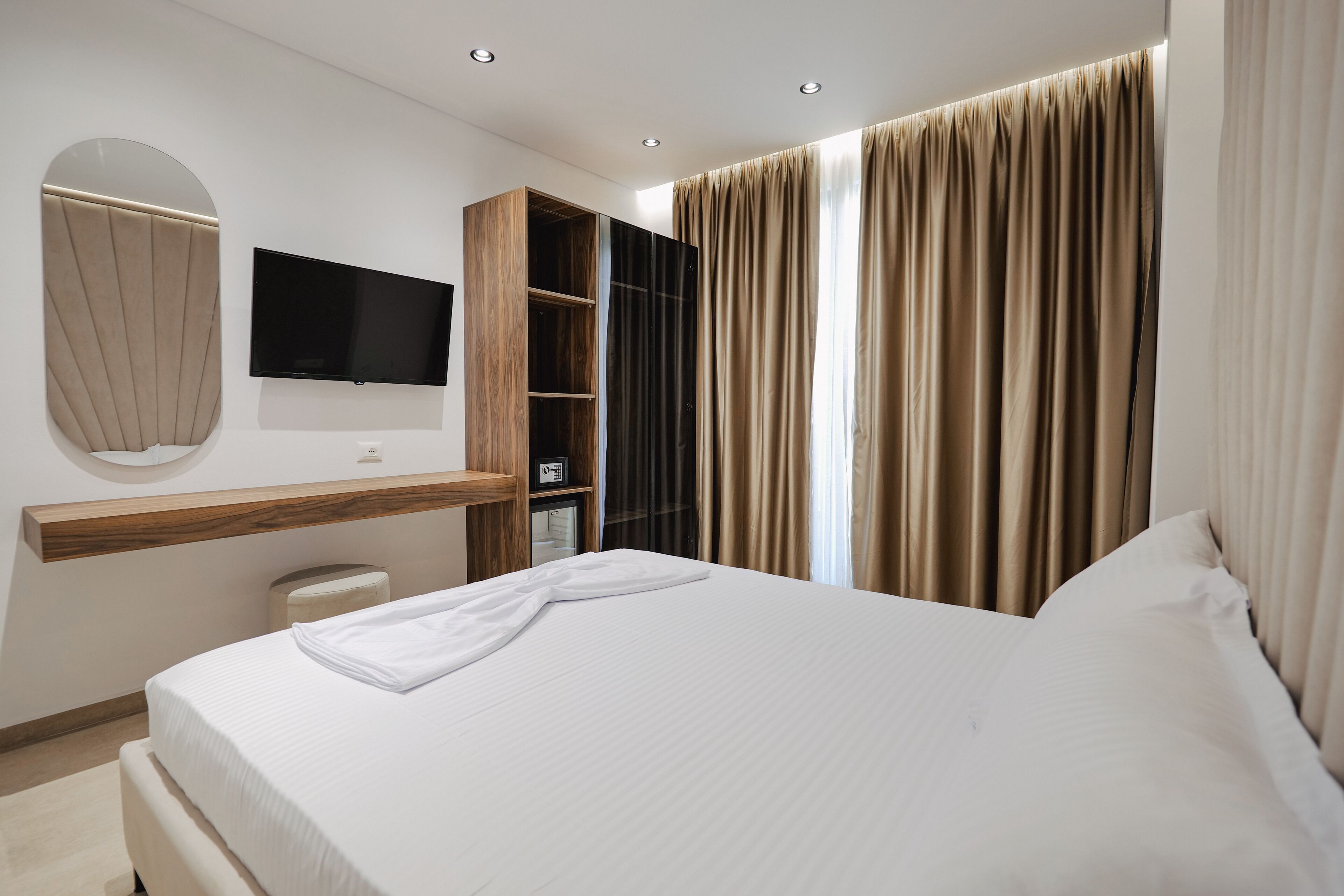 A modern hotel room with a large bed, a flat-screen TV mounted on the wall, a wardrobe, and a window with curtains.