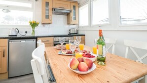 A modern kitchen with wooden cabinets, a dishwasher, and a dining table set with fruits, glasses, and a bottle. - Wagtails (Dartmouth)