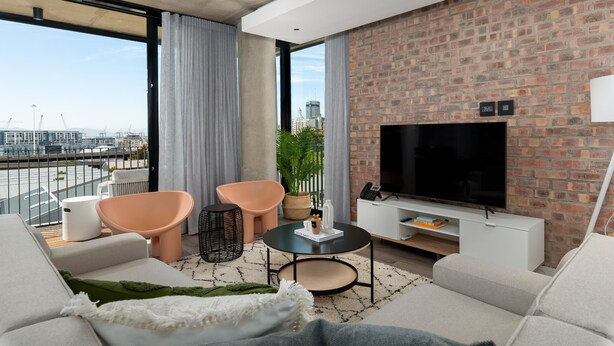 A modern living room with a large TV, comfortable seating, a brick accent wall, and a view of the city. - Trendy Signature Apartment l Wifi l Balcony (Cape Town)