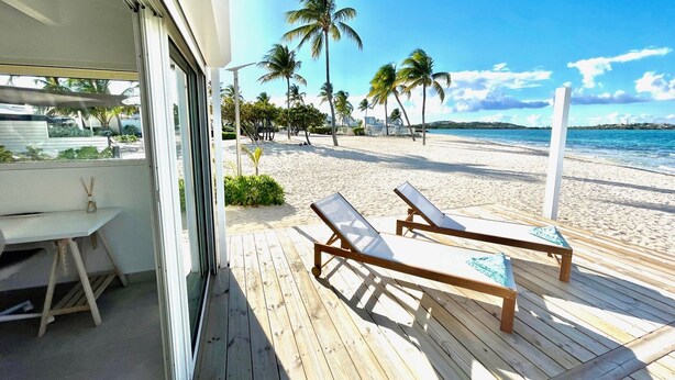 Eine Holzterrasse mit zwei Liegestühlen und einem Tisch, mit Blick auf einen Sandstrand mit Palmen und einem klaren, blauen Himmel.