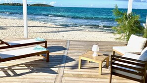 Eine Holzterrasse mit Liegestühlen und einem Tisch mit Blick auf einen Strand und klares blaues Wasser sowie eine entfernte Insel.