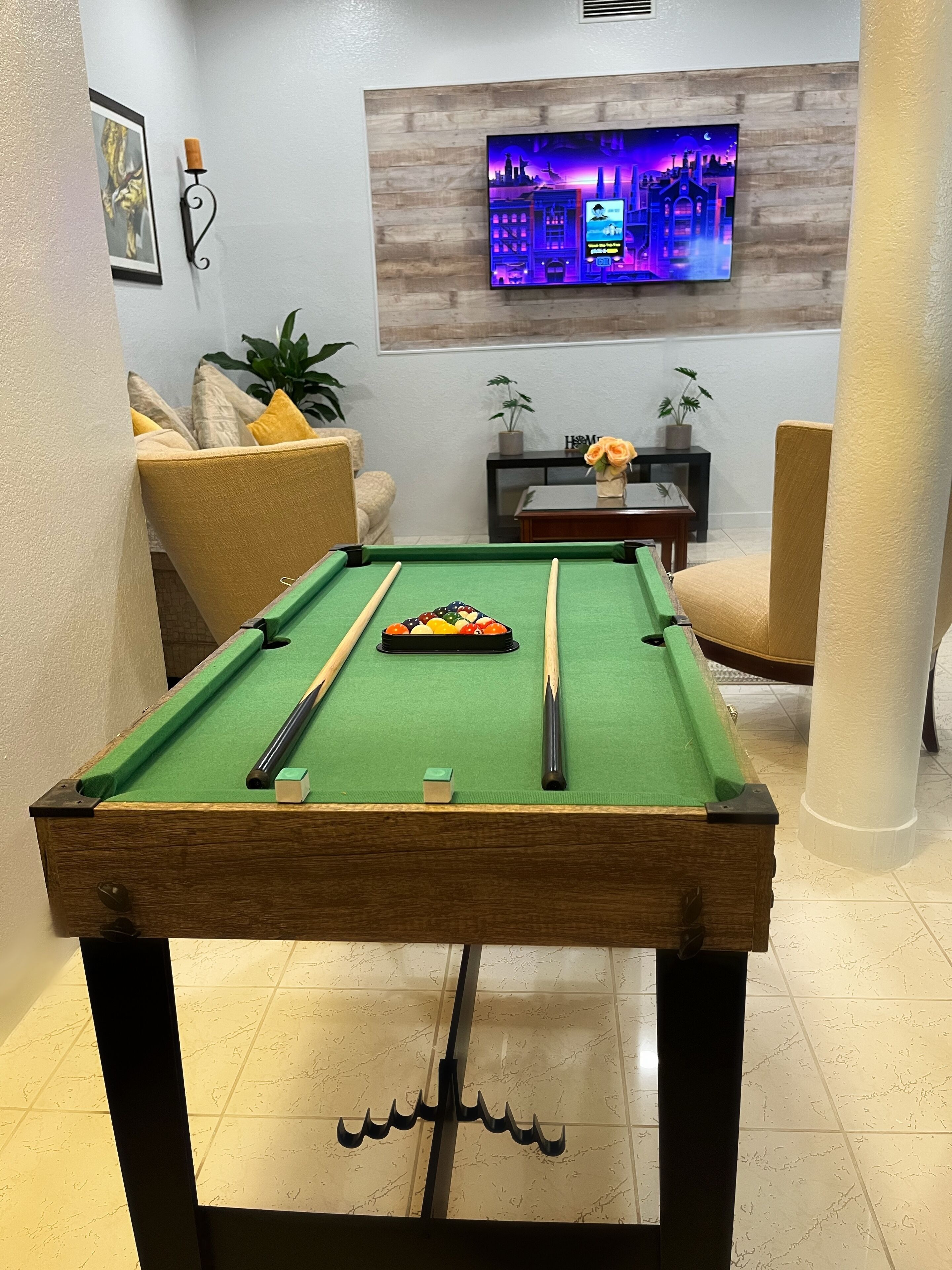 Une table de billard recouverte d’un drap de feutre vert, entourée d’un fauteuil beige, d’une petite table sur laquelle est posée une plante en pot et d’un téléviseur fixé au mur qui diffuse une vue sur la ville.