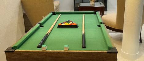 Une table de billard recouverte d’un drap de feutre vert, entourée d’un fauteuil beige, d’une petite table sur laquelle est posée une plante en pot et d’un téléviseur fixé au mur qui diffuse une vue sur la ville.