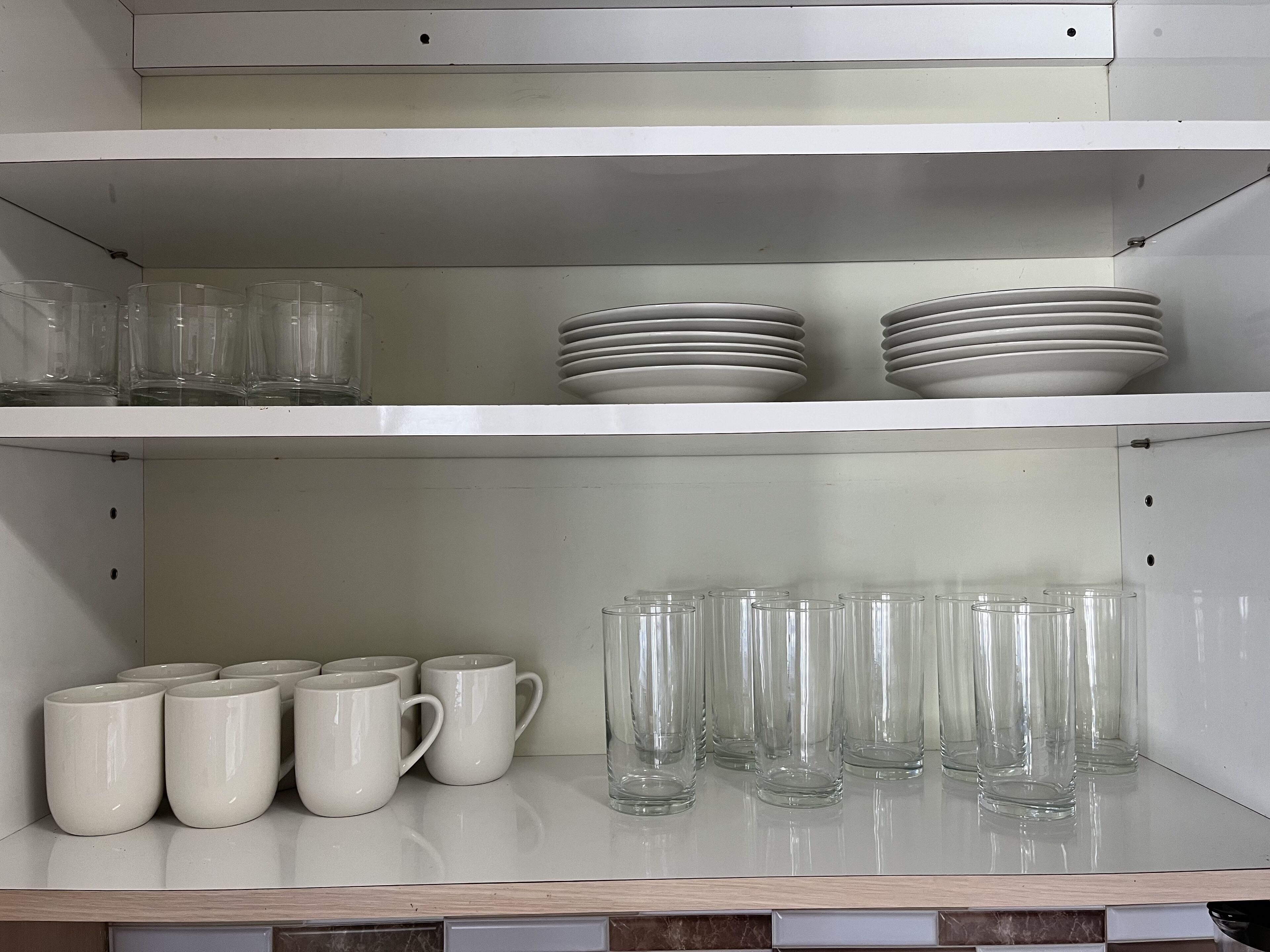 A kitchen shelf with clear glass mugs, white ceramic mugs, and stacked white plates.