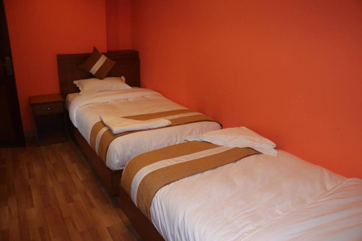 A room with a wooden floor, a single bed with white bedding and a brown accent, and a nightstand with a lamp.