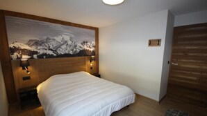 A hotel room with a large bed, wooden headboard, a mountain landscape painting, two wall-mounted lamps, and a wooden door. - 2nd floor, balcony, tv, 59m², Les Saisies (Hauteluce)