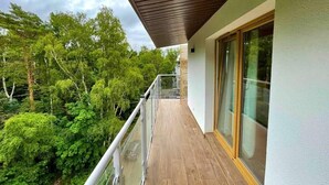 Un balcón con piso de madera, barandas de vidrio y vista a árboles verdes y frondosos.