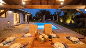 An outdoor dining area with a wooden table set for a meal, surrounded by chairs, and a pool in the background. - Modern villa with private pool - 120 m from Verde Blue beach (Μπαρμπάτι)