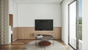 A modern living room with a flat-screen TV mounted on a wooden wall, a round coffee table with a book, and a large window offering a view of the outdoors.
