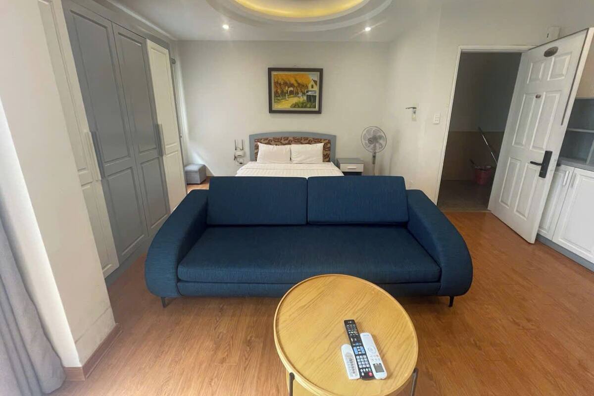 Une pièce avec un canapé bleu, une table basse en bois sur laquelle se trouve une télécommande, un lit avec une literie blanche, un tableau accroché au mur et un luminaire au plafond.