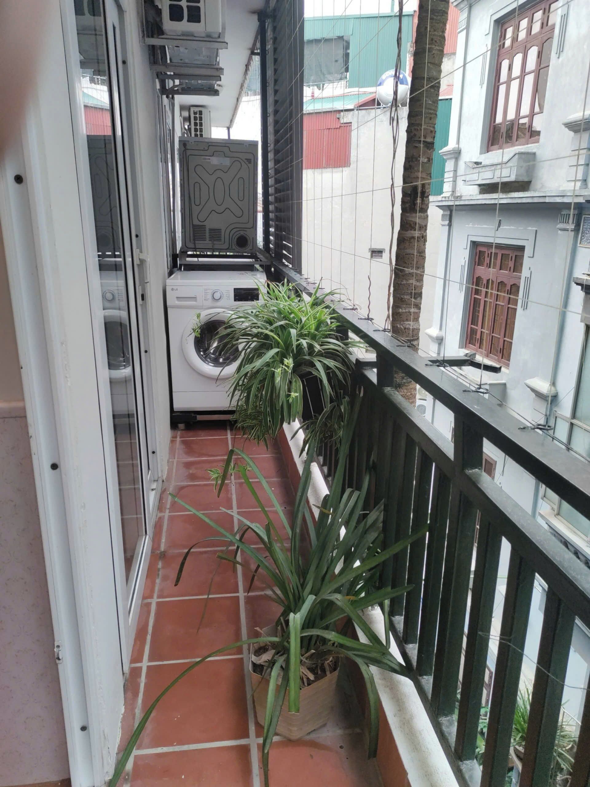 A narrow balcony with a washing machine, a potted plant, and a view of neighboring buildings.